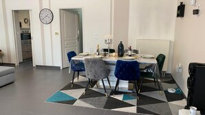 Un coin repas avec une table dressée pour quatre personnes, une horloge au mur et une cuisine en arrière-plan.