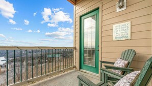 A balcony with a view of a river, a green door, and two Adirondack chairs with cushions.