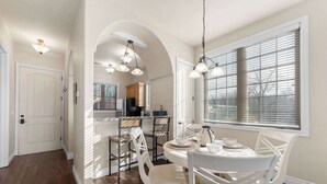 A dining area with a round table, white chairs, and a kitchen in the background.