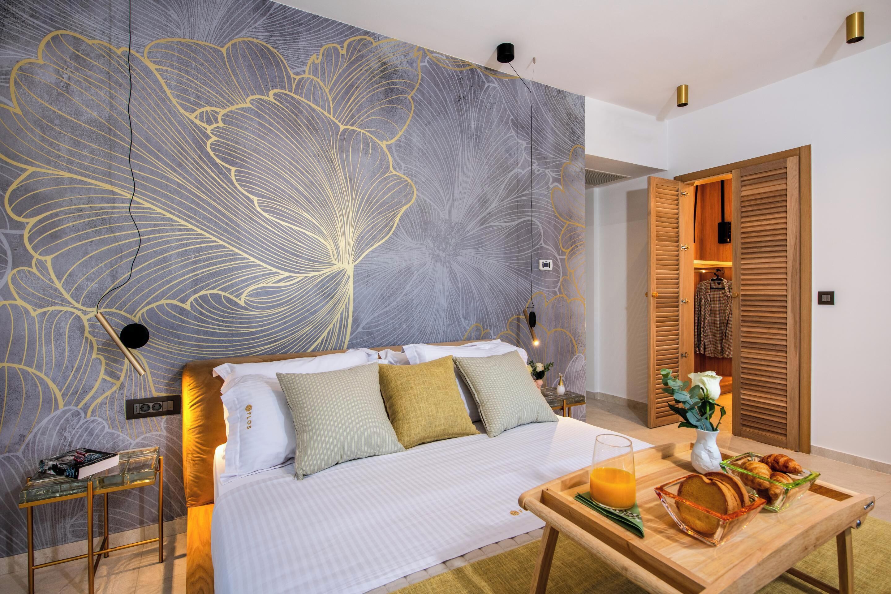 A modern bedroom with a large floral wall mural, a neatly made bed with pillows, a bedside table with a book, and a breakfast tray on a wooden table.