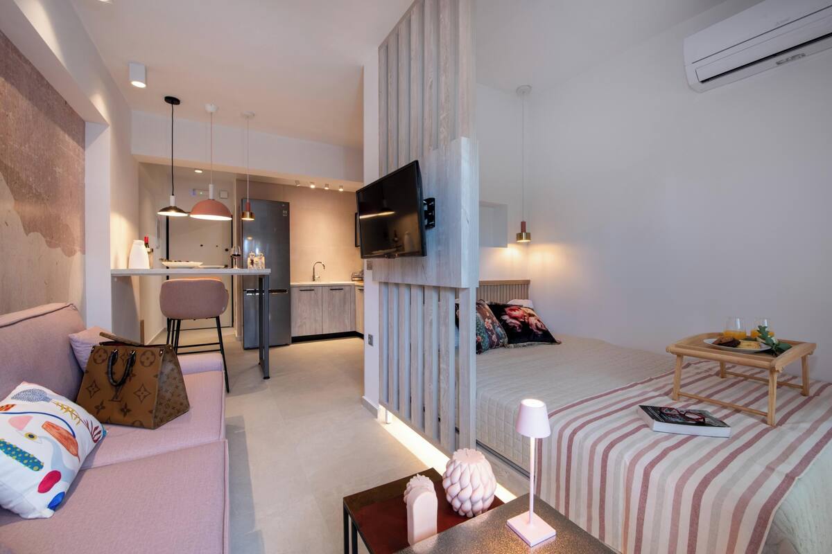 A modern hotel room with a bed, a sofa, a small table, a TV mounted on the wall, and a kitchenette area.