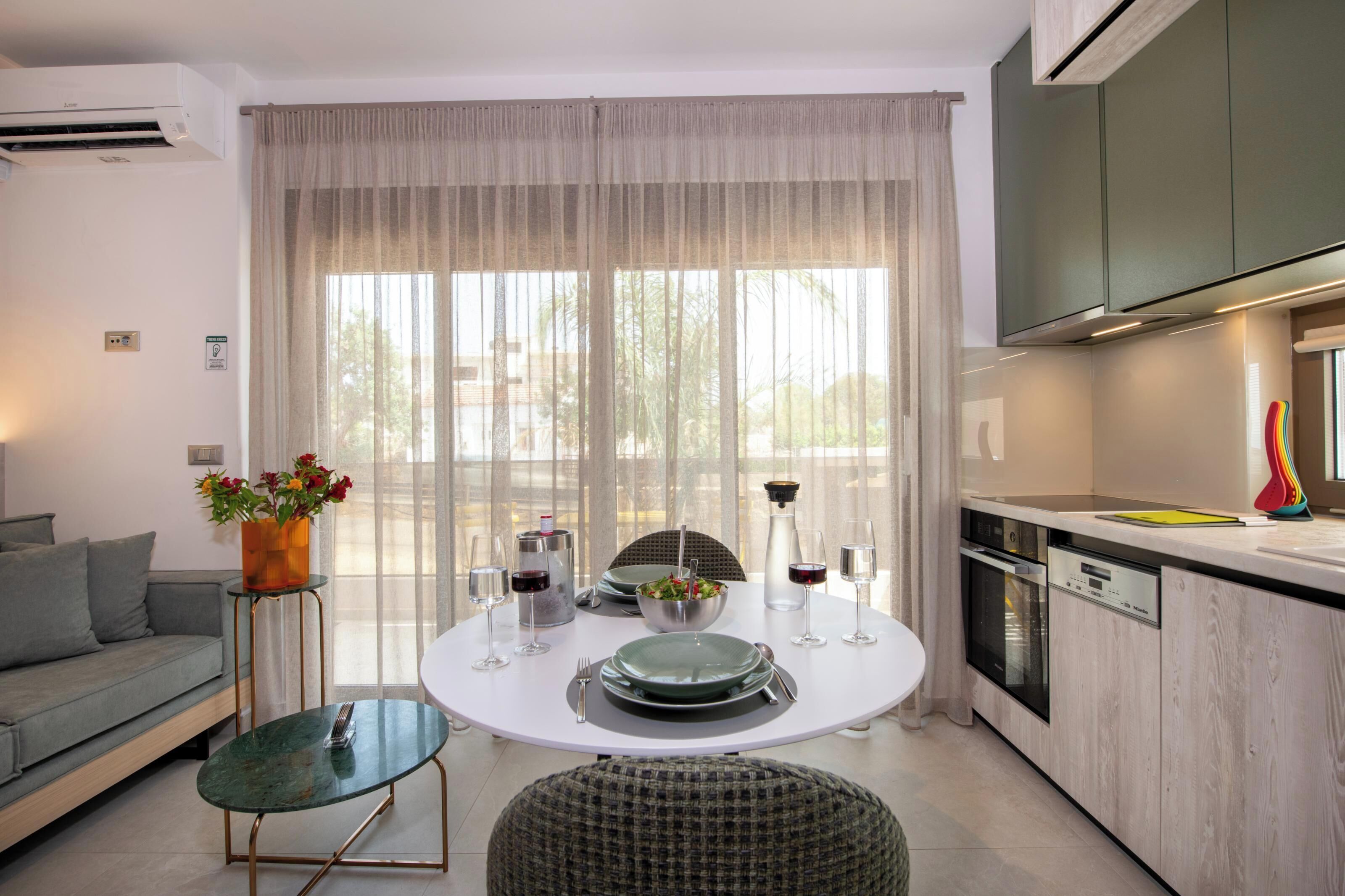 A modern kitchen with a dining table set for two, a sofa, and a window with sheer curtains.