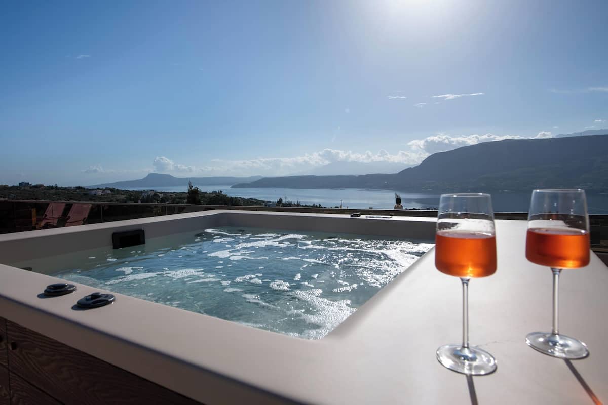 A hot tub with two glasses of wine on a table, overlooking a body of water and mountains.