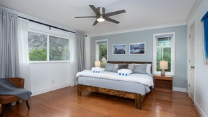 A bedroom with a wooden bed, a ceiling fan, two bedside lamps, and a view of trees through the windows. - New! Big Sky Cabin at Yosemite w/ VIEWS, hot tub, firepit, and more! (Oakhurst)