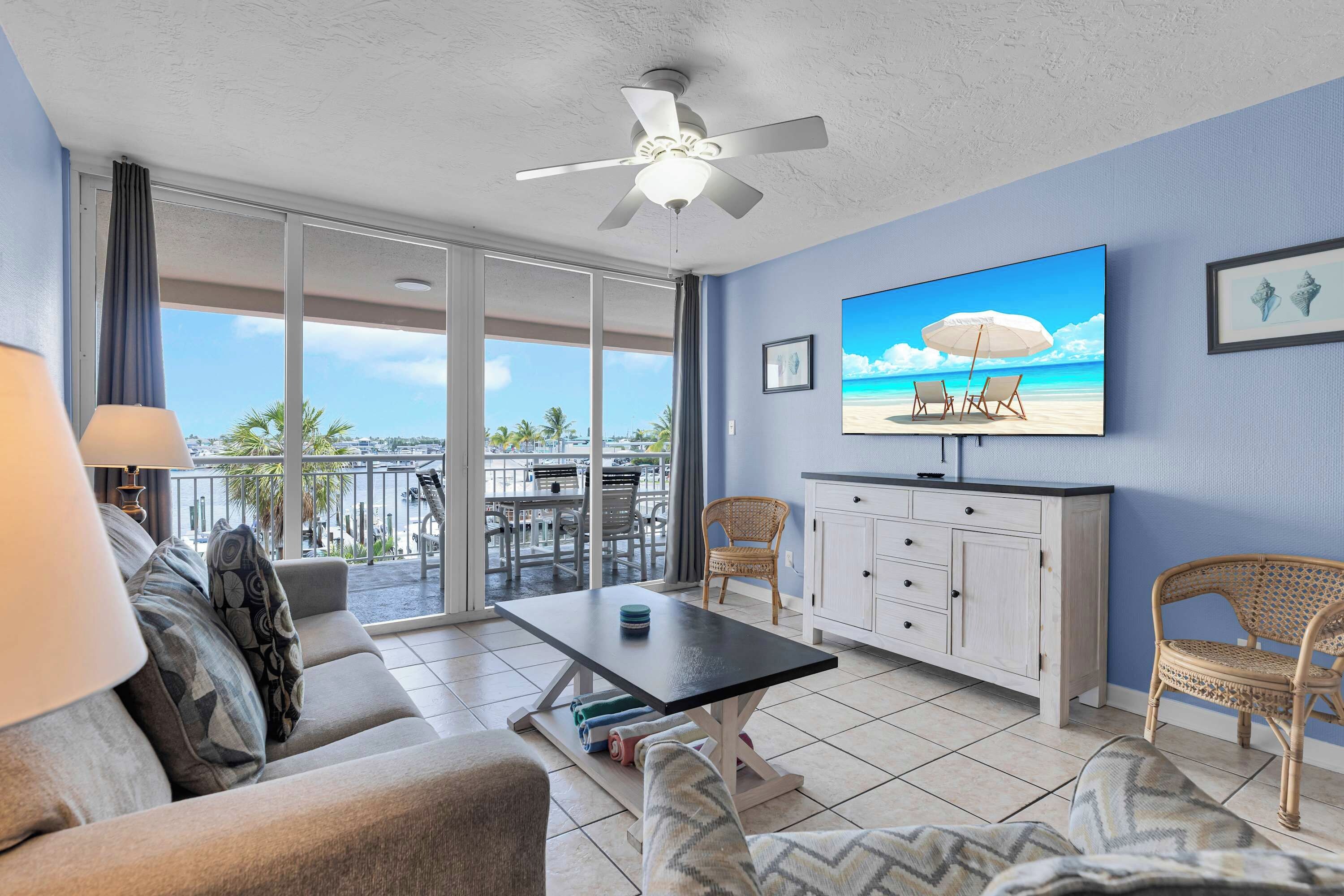 A living room with a TV, sofa, and chairs, a balcony with a view, and a ceiling fan.