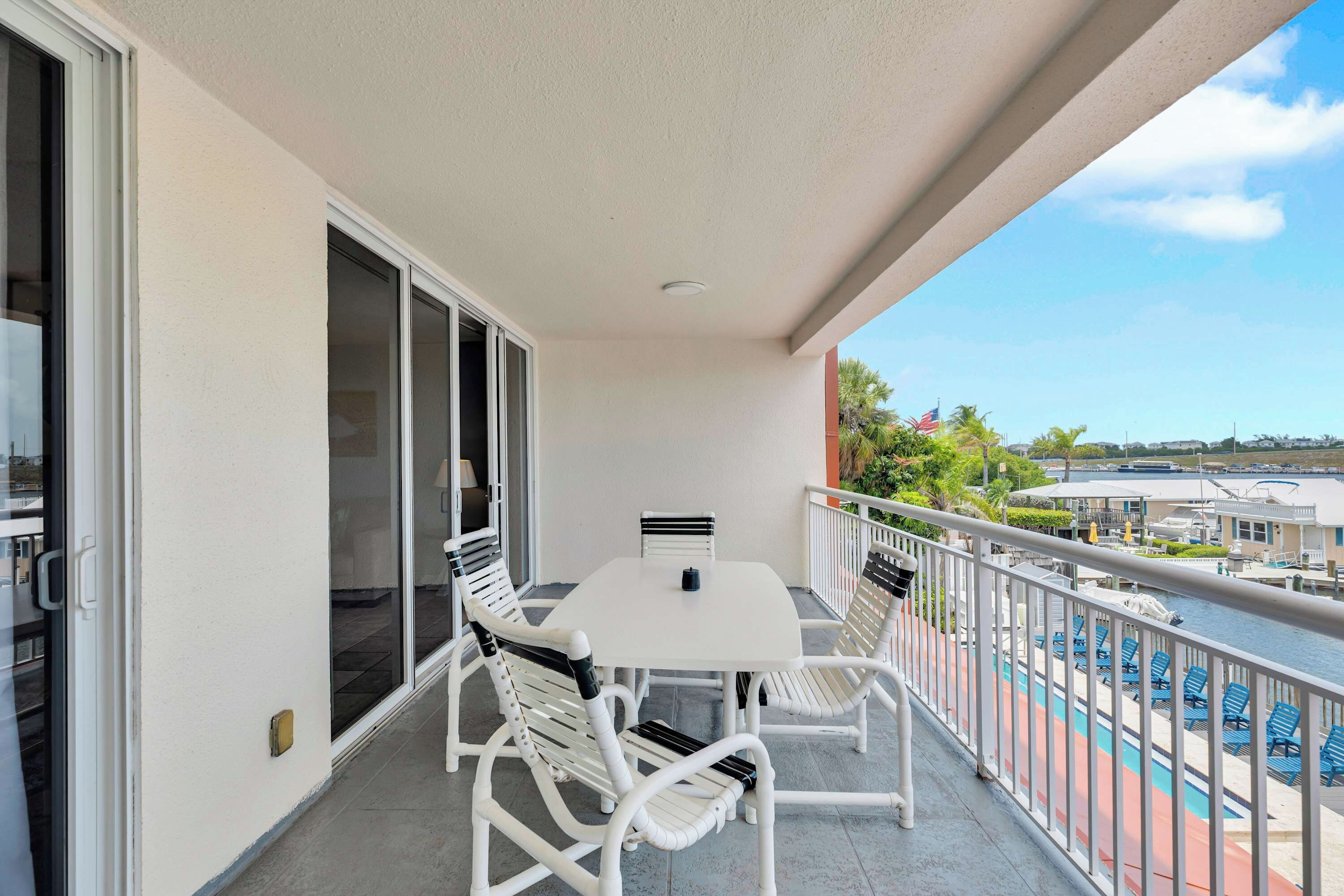 A balcony with a white table and chairs, a view of a swimming pool, and a building with a flag.