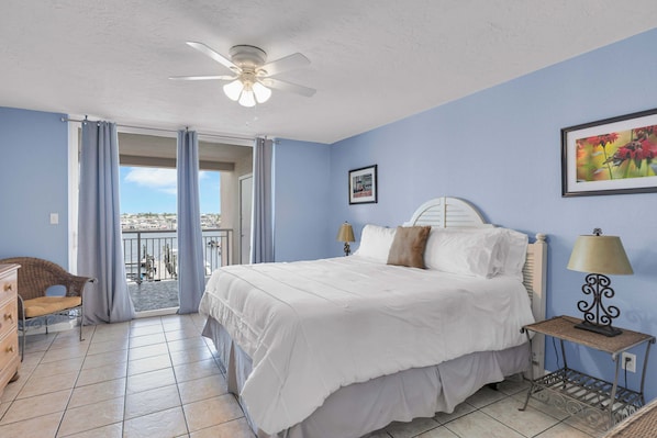 A bedroom with a bed, a ceiling fan, a window with curtains, a chair, and a small table with a lamp.