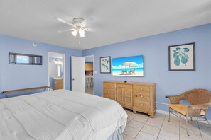 Un dormitorio con cama, cómoda, silla, ventilador de techo, televisor y puerta que da a un baño.