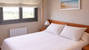 Una cama bien tendida con una colcha blanca, un cabecero de madera y una mesita de noche con una lámpara. Se ve una ventana con vistas al exterior y un cuadro en la pared.