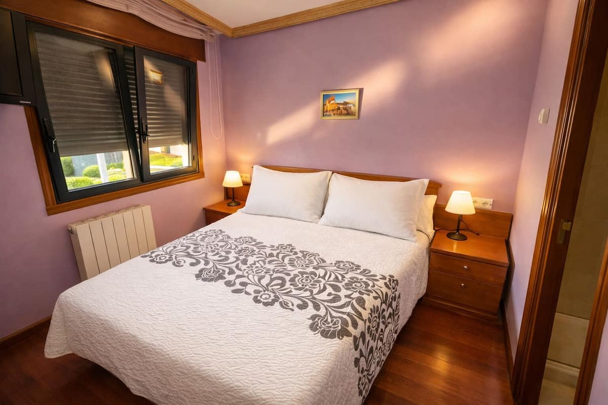A bedroom with a bed, two bedside tables with lamps, a radiator, and a window with blinds.