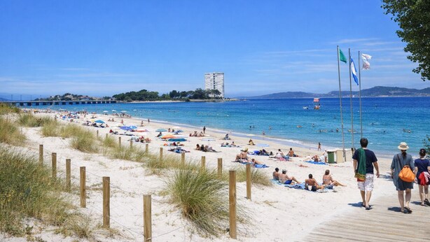 Una playa con gente tomando el sol y nadando, un paseo marítimo de madera y un edificio a lo lejos.