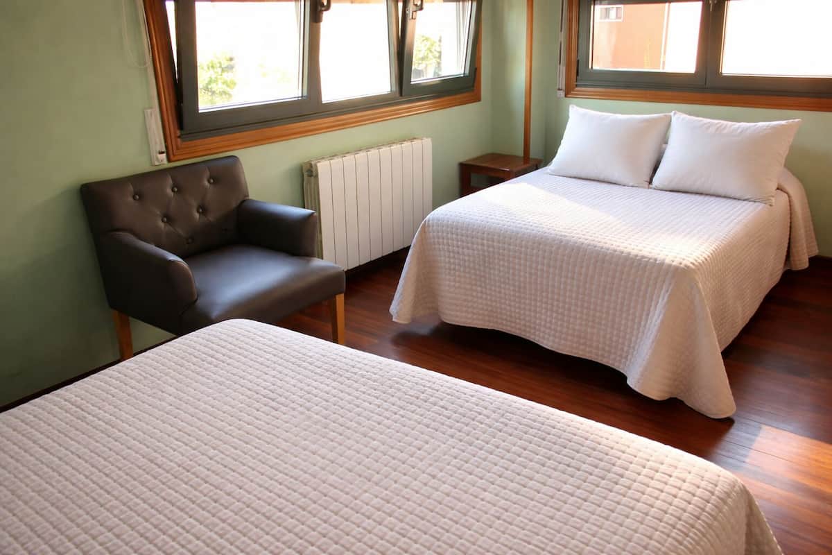 A room with two beds, a chair, a radiator, and windows.
