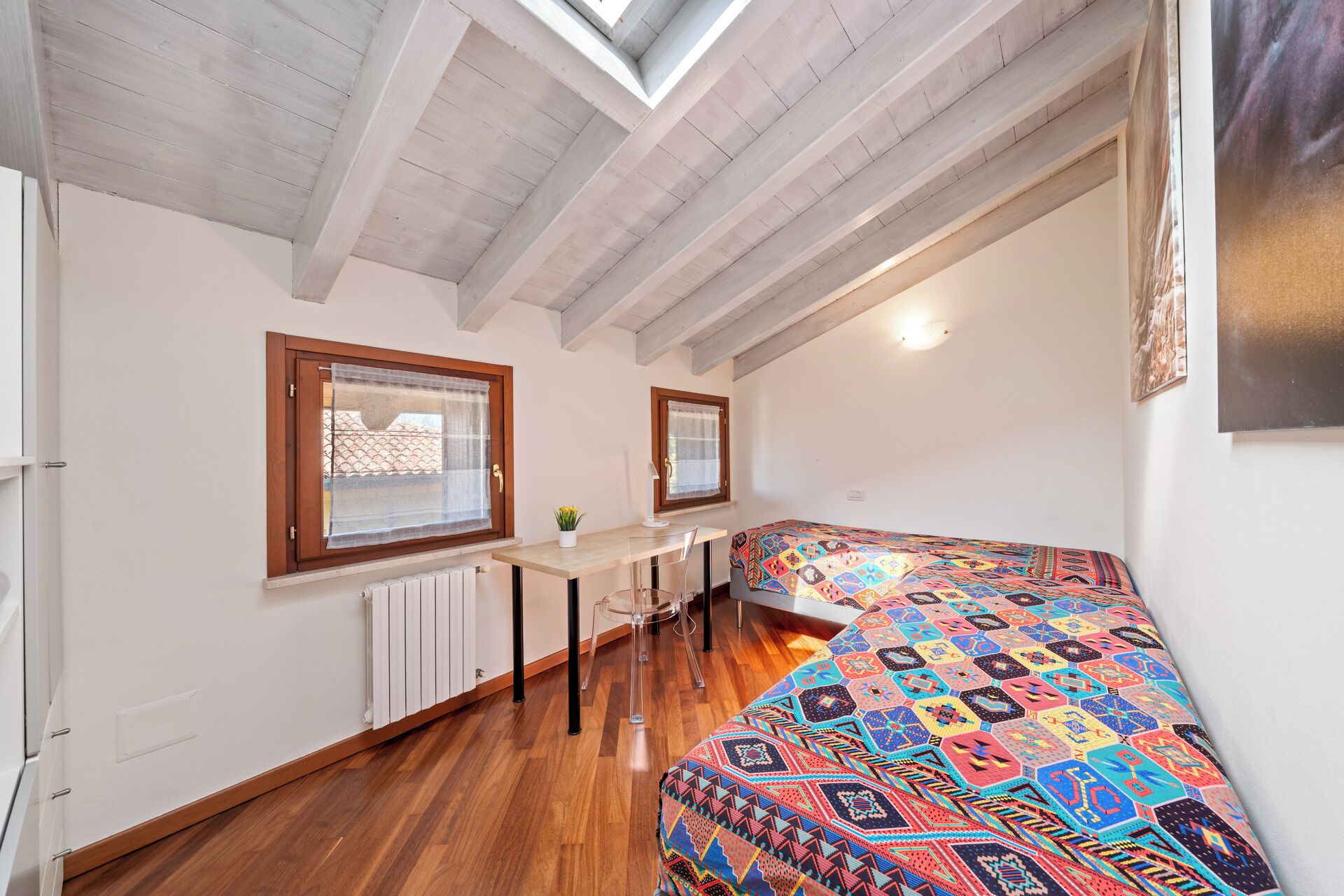 A small room with a sloped ceiling, a bed with a colorful patterned bedspread, a desk with a chair, and a window with a curtain.