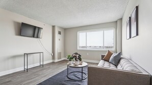 A modern living room with a flat-screen TV mounted on the wall, a round coffee table with plants, a sofa with cushions, and a large window providing a view. - Legacy at Imperial Village 1014 I 2 Bed 2 Bath (Ric hmond)