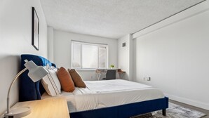 Un dormitori modern amb un llit blau, roba de llit blanca, escriptori amb cadira i finestra amb persianes.