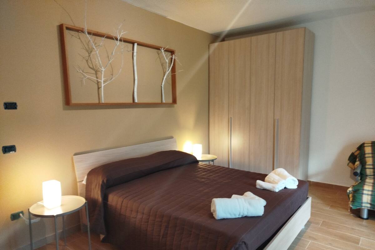A neatly made bed with a brown bedspread and white towels, a bedside table with a lamp, a wardrobe, and a chair with a plaid blanket.