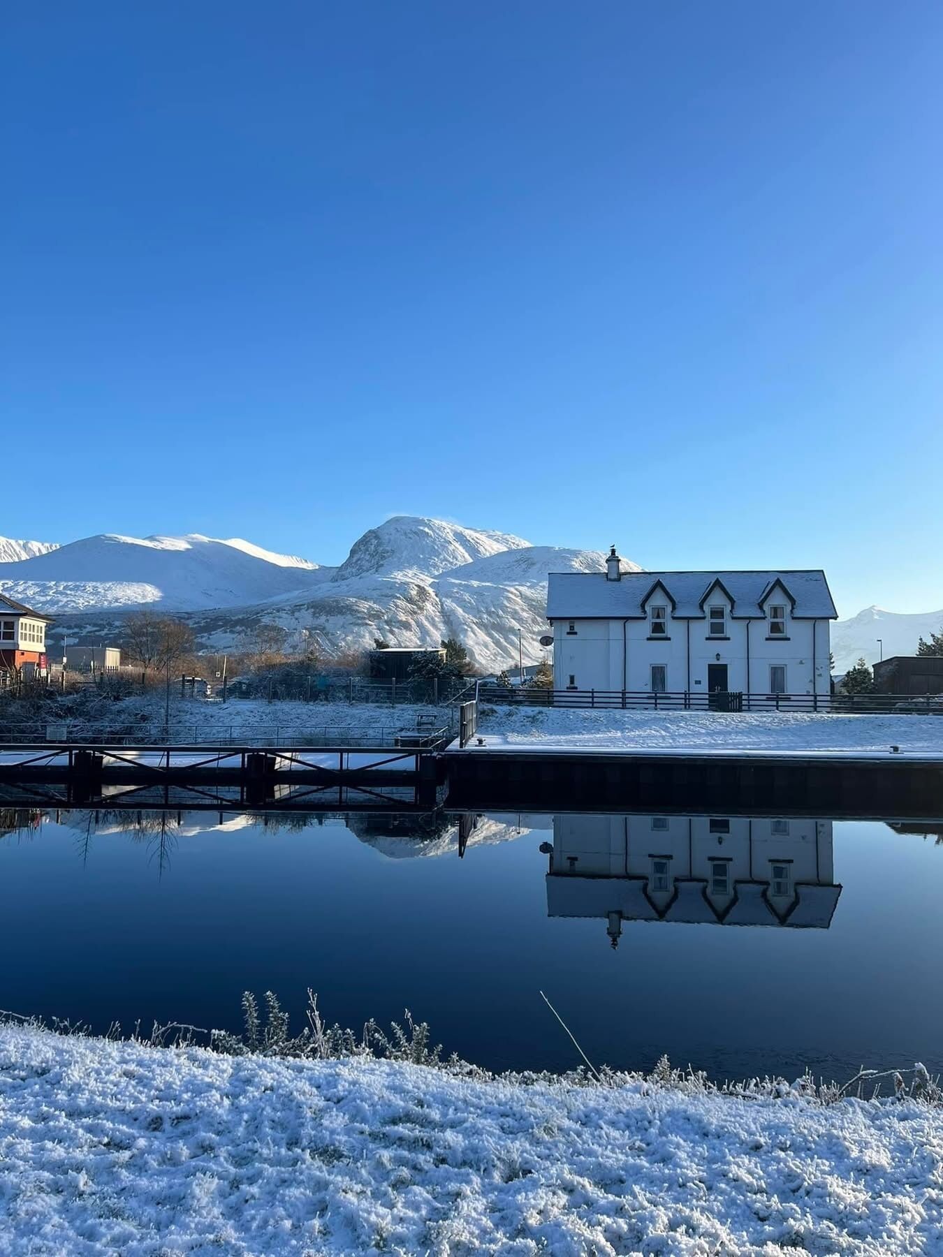 A white building with a symmetrical facade, a dock extending into a calm body of water, and snow-covered mountains in the background.