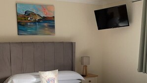 Una cama bien hecha con cabecera gris, un cuadro de un paisaje sobre ella, una mesita de noche con una lámpara y un televisor montado en la pared.