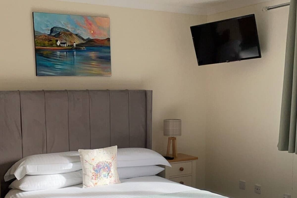A neatly made bed with a grey headboard, a painting of a landscape above it, a bedside table with a lamp, and a TV mounted on the wall.