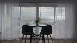 Una habitación con una mesa redonda y dos sillas, una planta en maceta sobre la mesa, y vistas amplias a través de ventanas con persianas.