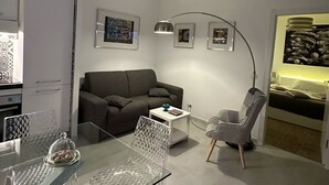 A modern living room with a glass dining table, a grey sofa, a grey armchair, and a white coffee table. There are framed pictures on the wall and a ceiling light fixture. - Isoco Terrace (Taormina)