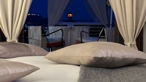 A bed with beige and gray bedding, a view of the cityscape at dusk, and a table with chairs in the background. - Isoco Terrace (Taormina)