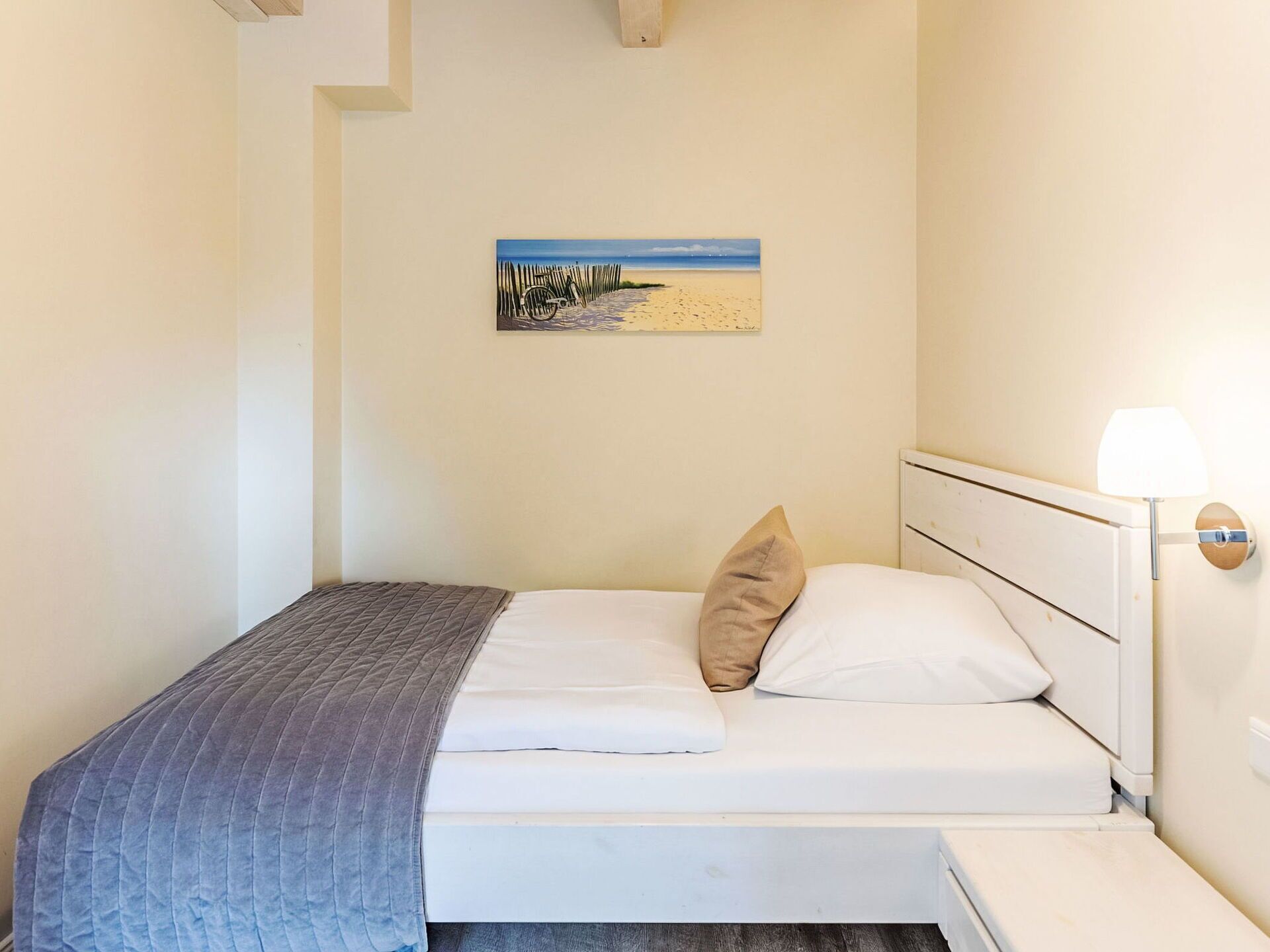 A single bed with a white headboard, a beige pillow, and a blue blanket. There is a painting of a beach scene on the wall above the bed.