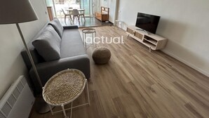A modern living room with a grey sofa, a TV on a wooden stand, and a dining area visible through glass doors. - Charming apartment with pool near the beach in Platja d'Aro (Platja d'Aro)