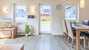 A modern, well-lit room with a wooden ceiling, a dining table with chairs, a TV, and a large window with a view of a house. - Lovely apartment in villa for 2 people with WIFI, TV and terrace (Dagebüll)