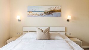 A neatly made bed with white linens and a beige pillow, a painting of a beach scene on the wall, and two wall-mounted lamps on either side of the bed. - Lovely apartment in villa for 2 people with WIFI, TV and terrace (Dagebüll)