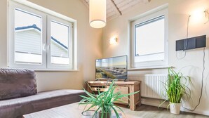 A modern living room with a sofa, a wooden coffee table with decorative items, a TV on a wooden stand, and large windows with blinds. - Lovely apartment in villa for 2 people with WIFI, TV and terrace (Dagebüll)