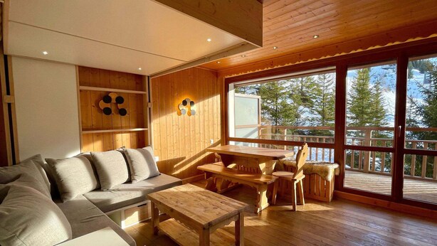 Ambiente moderno con soffitto in legno, divano, tavolo da pranzo e vista su un paesaggio innevato attraverso ampie finestre.