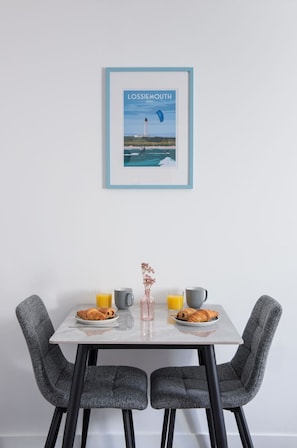 Uma área de refeições com uma mesa posta para o café da manhã, incluindo croissants e suco de laranja, e uma moldura com a imagem de um farol na parede.