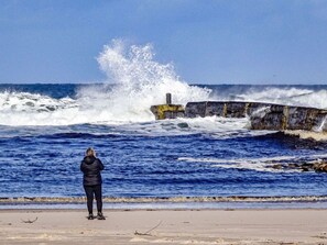 A person standing on a sandy beach facing a rocky breakwater with waves crashing against it.