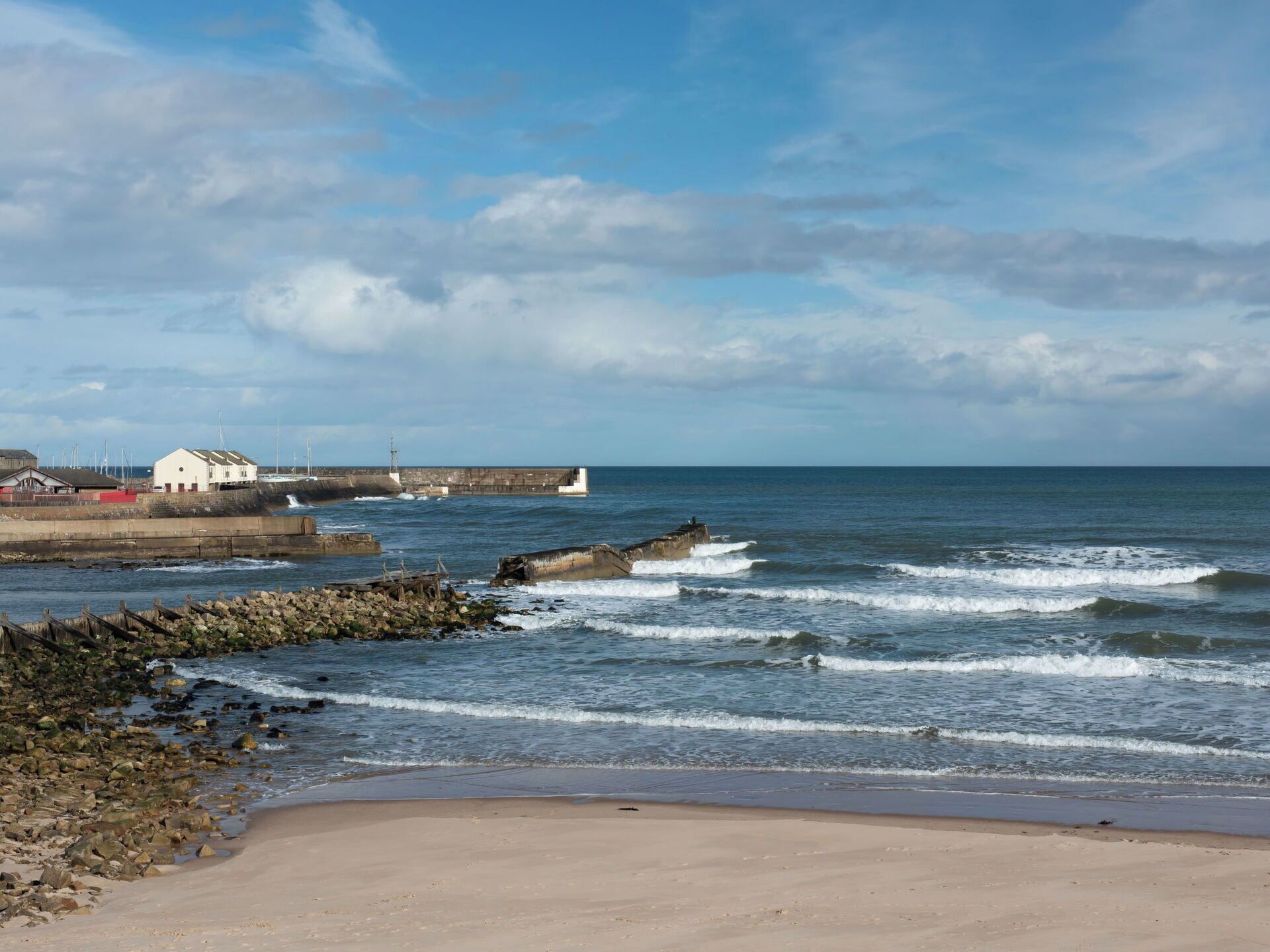 A coastal scene with a sandy beach, rocky shoreline, and a breakwater extending into the sea.