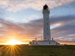 A tall white lighthouse with a balcony, set against a sunset sky.