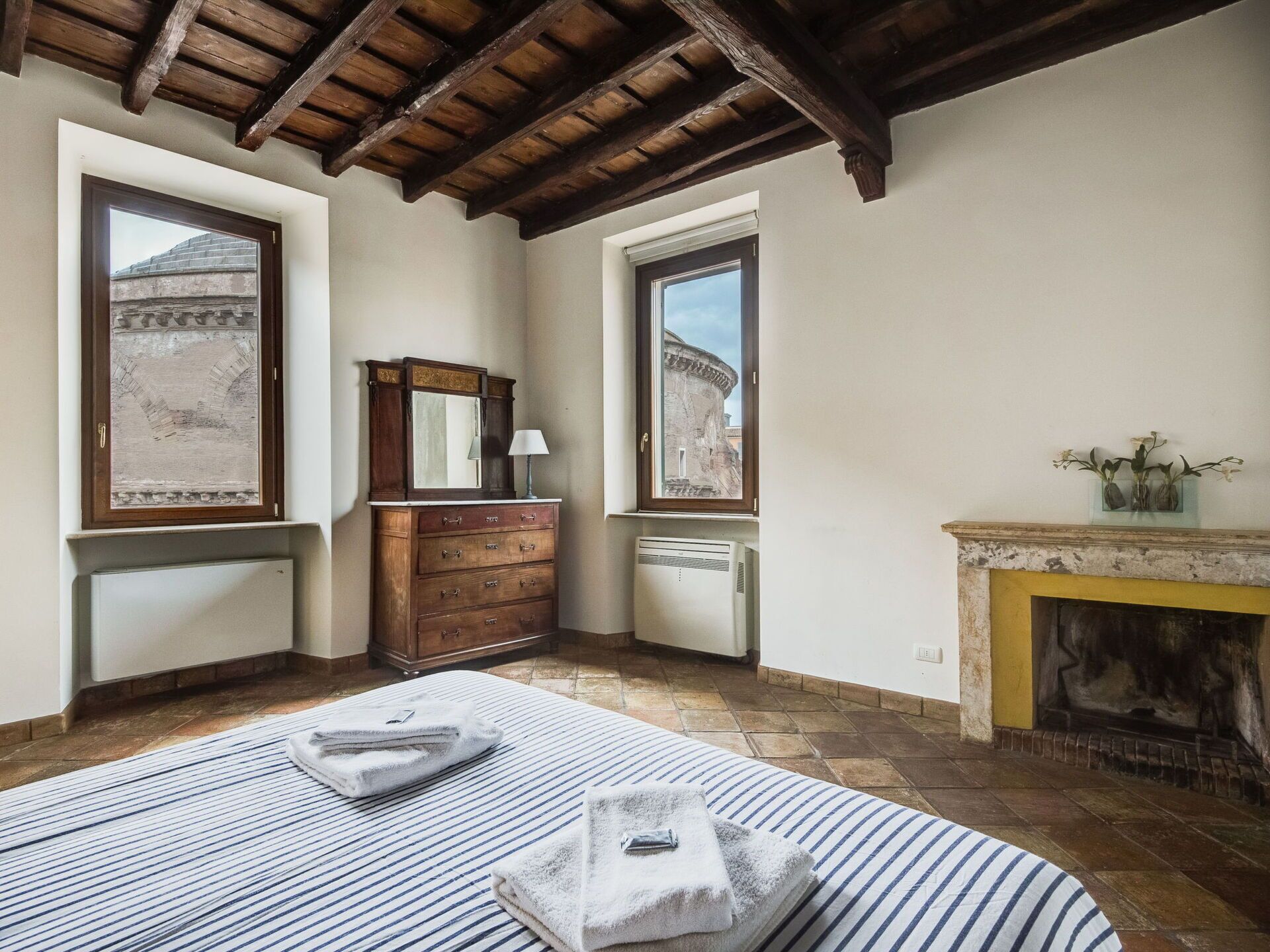 Une chambre avec un plafond en bois, une cheminée, une grande fenêtre donnant sur la ville, une commode avec des tiroirs et un lit avec une literie à rayures.