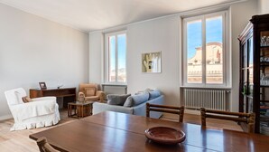 Ein großzügiges Wohnzimmer mit einem Holzes Esstisch, einer Couch und einem Sessel. Große Fenster bieten Blick auf Gebäude.