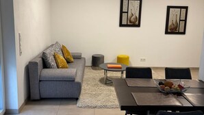 Un salon moderne avec un canapé gris, des coussins jaunes, une table à manger avec des chaises et deux œuvres d’art encadrées accrochées au mur.