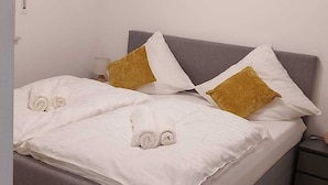 A neatly made bed with white linens and two pillows, a bedside table with a lamp, and a chair in a room with wooden flooring and a grey headboard. - Beautiful apartment in a quiet neighborhood (Maxhütte-Haidhof)