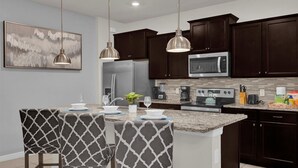 A modern kitchen with dark wood cabinets, stainless steel appliances, and a granite countertop. There are two pendant lights, a refrigerator, a microwave, a stove, and a coffee maker.