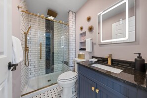 Un baño con ducha envuelta en vidrio, inodoro blanco, tocador con encimera negra y espejo con marco iluminado.
