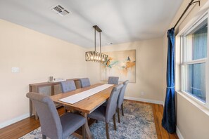 Un coin repas avec une table en bois, des chaises à revêtement gris, un luminaire suspendu et un grand tableau abstrait accroché au mur.