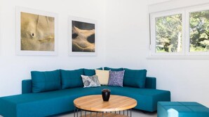 Un sofá azul verdoso con una mesa redonda de madera en una sala de estar moderna, con dos obras de arte enmarcadas en la pared y una ventana que ofrece vistas a árboles.