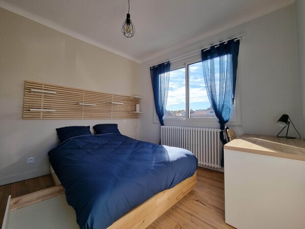 A bedroom with a bed, a bedside table, a radiator, a window with curtains, a hanging light fixture, and a wall-mounted shelf.