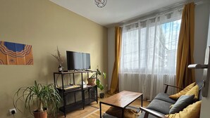 Ein Wohnzimmer mit einer Couch, einem Couchtisch, einem Fernseher auf einem Schrank und einem Fenster mit Vorhängen.