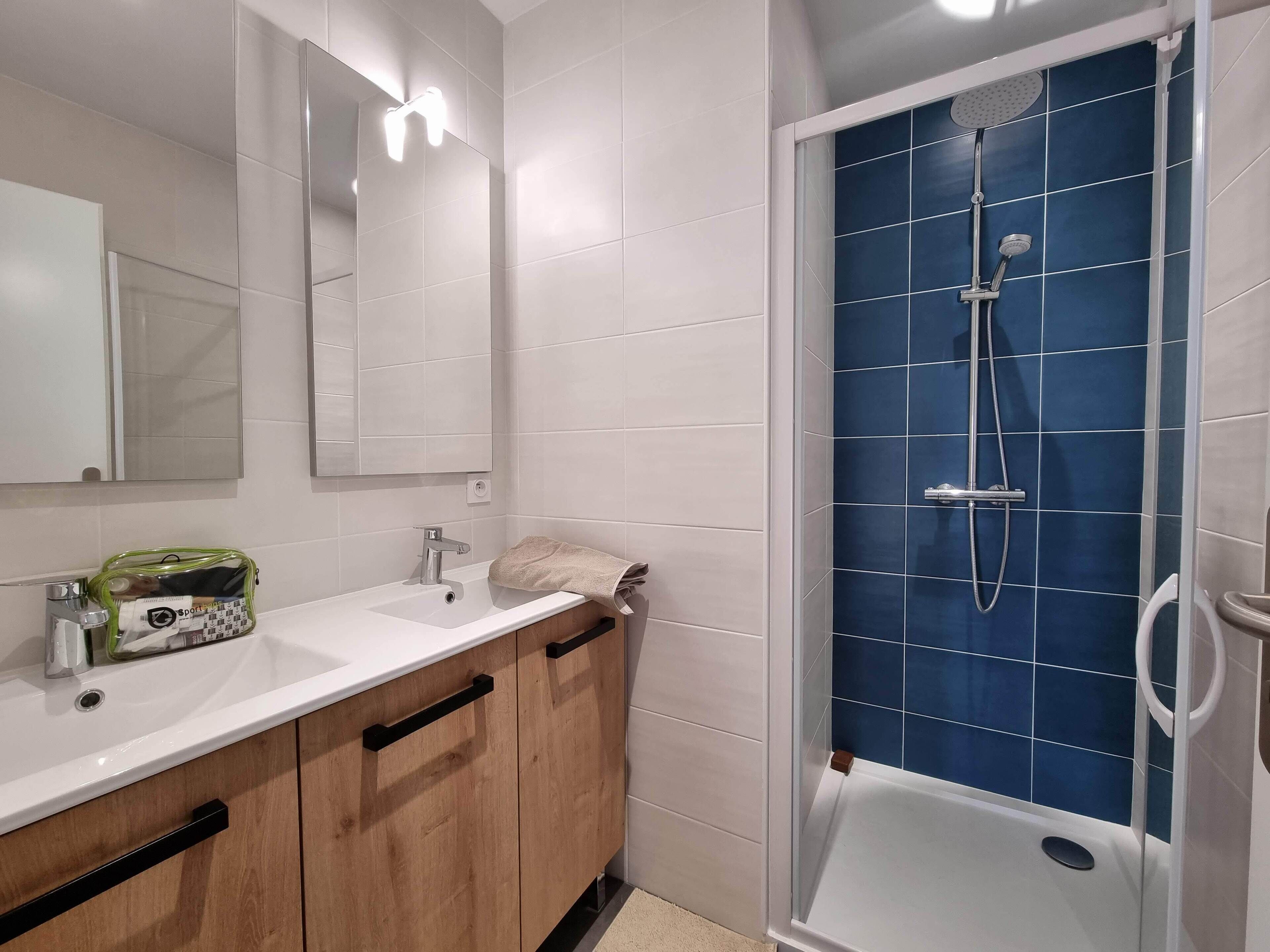 Un bagno moderno con lavandino bianco, mobili in legno, uno specchio e zona doccia rivestita di piastrelle blu.