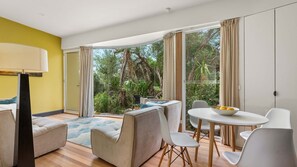 A modern living room with a sofa, chairs, a dining table, and a lamp. There is a large window with curtains, and a view of greenery outside. - Sea Glass Studio Inverloch by the sea (Inverloch)