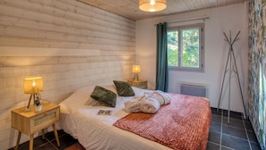 Ett sovrum med ett träpanelat tak, en säng med ett rött överkast, två kuddar, en sänglampa och ett fönster med gardiner.