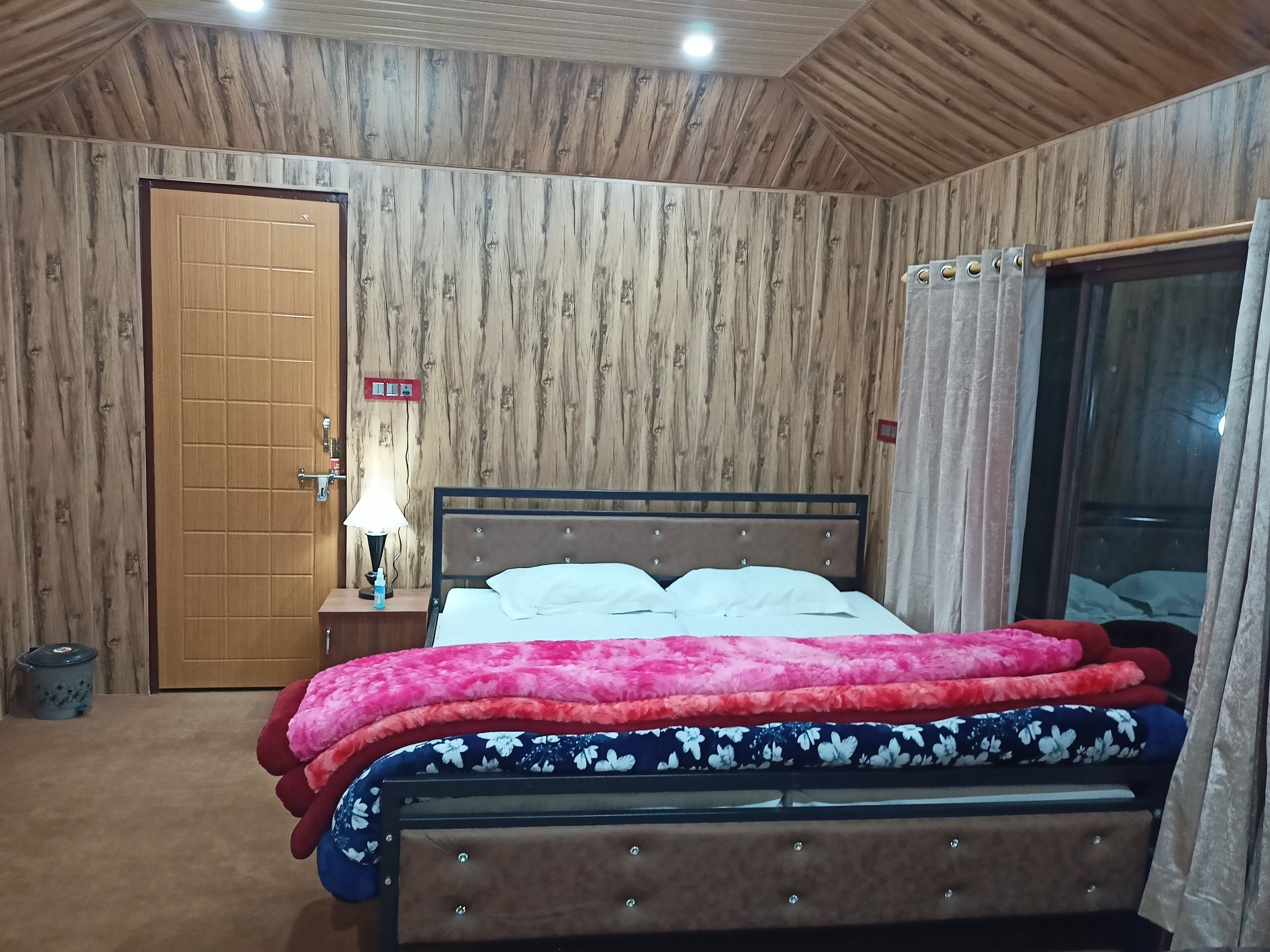 Un dormitorio con paneles de madera en la pared, una cama con varias almohadas, una mesita de noche con lámpara y un ventanal con cortinas.